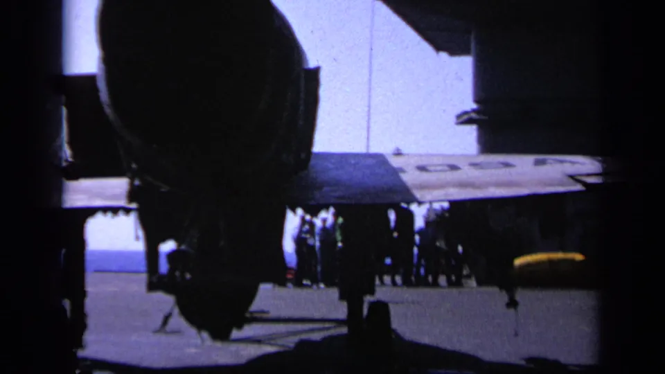 Greece 1963 8mm frame showing aircraft landing gear, a wing, and distant personnel gathered on a carrier deck.