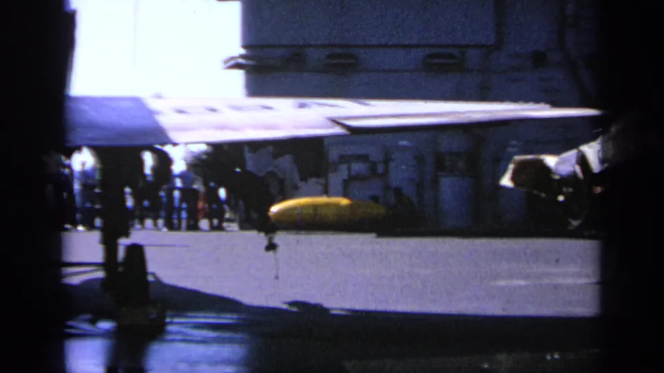 Greece 1963 frame showing a damaged aircraft on a carrier deck with personnel nearby and yellow equipment under the wing area.