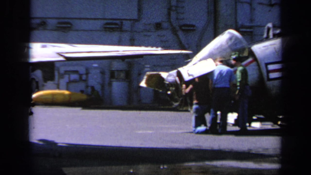 1963 Greece home-movie frame showing personnel gathered by the open nose area of a U.S. Navy jet on an aircraft carrier deck.