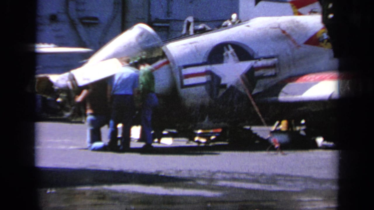 Personnel stand beside a white Navy jet with its canopy open.
