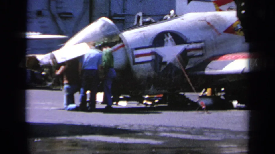 A U.S. Navy jet with star insignia and open canopy is serviced by personnel on a carrier deck in 1963 Greece footage.