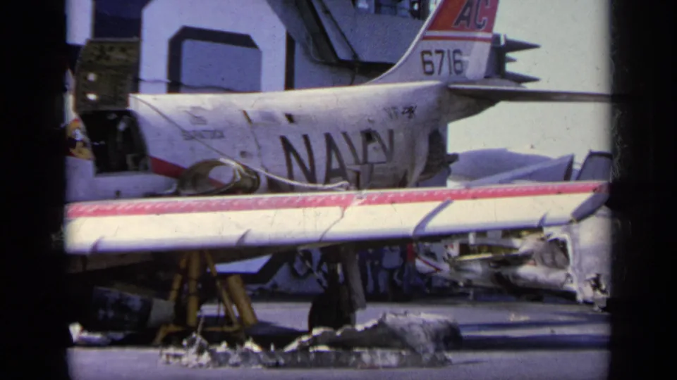 1963 Greece 8mm frame showing a damaged Navy aircraft marked 6716, torn wing sections, and debris on the tarmac.