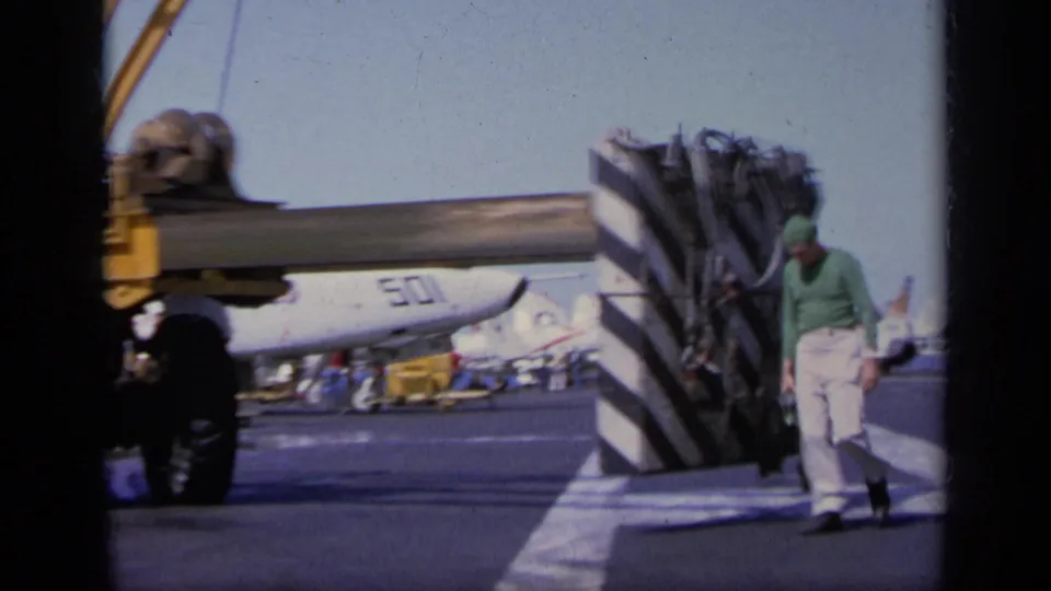 1963 Greece 8mm frame showing a worker near striped deck equipment, with aircraft and recovery gear visible in the background.