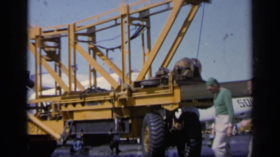 1963 Greece airfield frame with a man walking beside a large yellow aircraft handling structure and a white military jet marked 501 behind it.