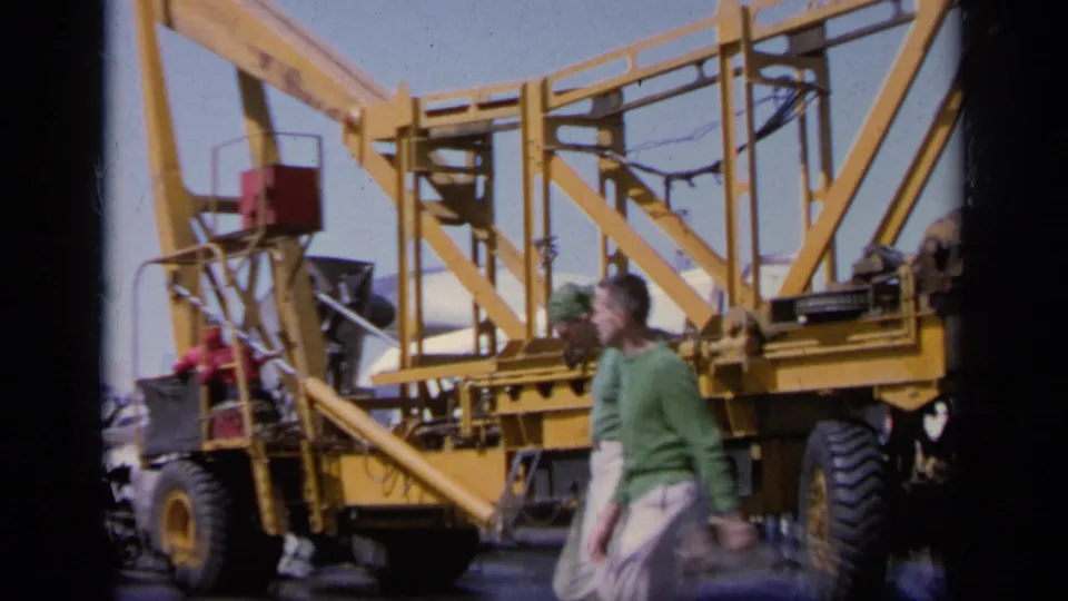 1963 Greece 8mm frame showing two men crossing an airfield in front of yellow wheeled aircraft equipment with ladders and railings.