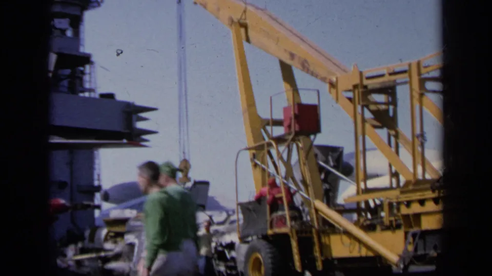 1963 Greece airfield frame with men walking beside yellow aircraft handling equipment, distant aircraft, and deck machinery in view.