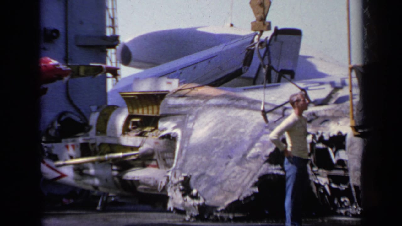 A crane line holds damaged aircraft wreckage beside ship structures and crew.