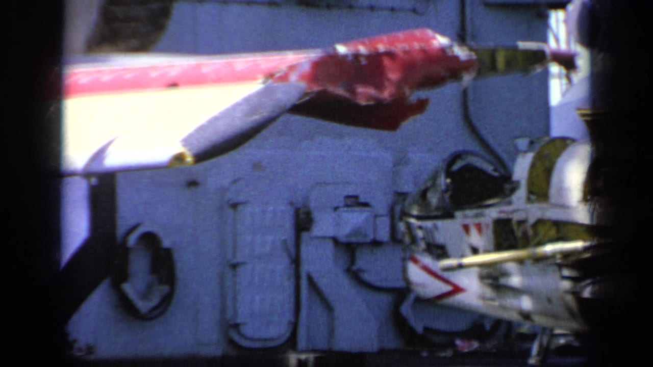 1963 8mm frame showing a close view of a U.S. Navy aircraft on the USS Saratoga, with wing markings and cockpit detail visible.