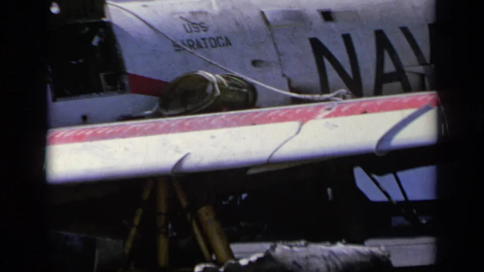 1963 8mm frame showing the aircraft side with large Navy lettering, USS Saratoga text, canopy detail, and red-and-white wing edge.