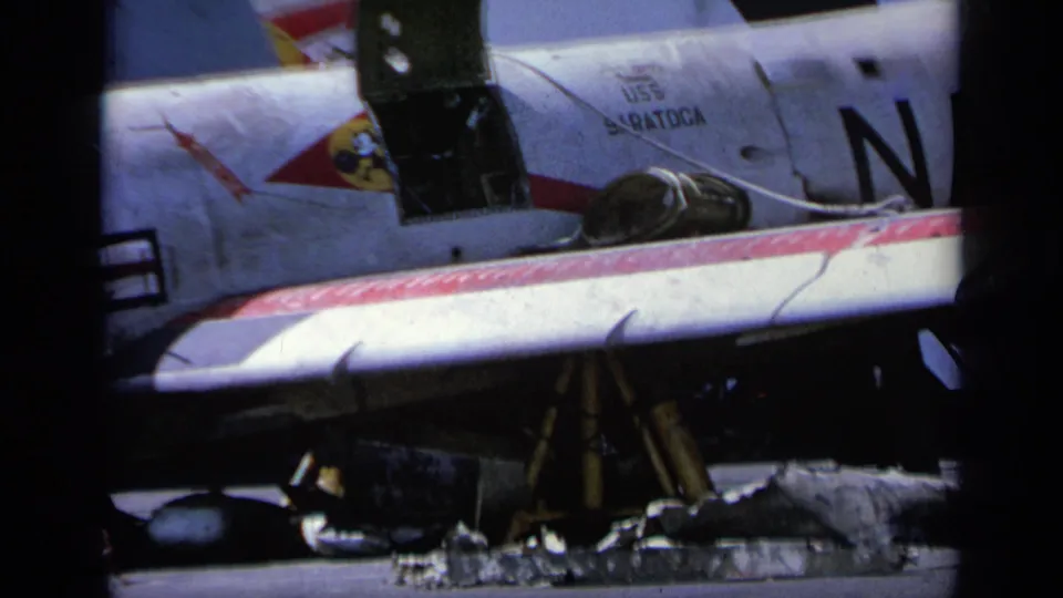 1963 8mm frame showing USS Saratoga text, a squadron-style insignia, cockpit canopy, and partial Navy lettering on the aircraft side.