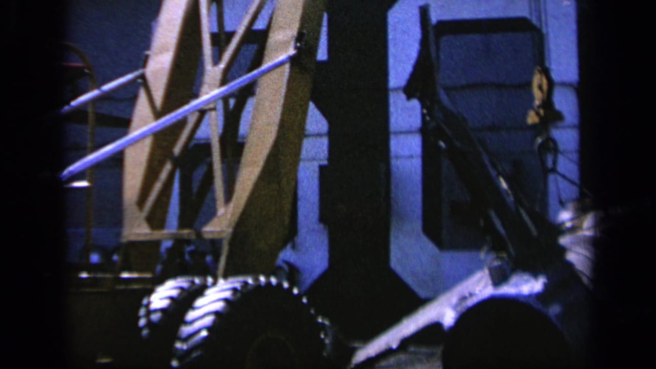 1963 Greece 8mm frame showing crane straps near a large cylindrical machine component in a blue-lit industrial interior.