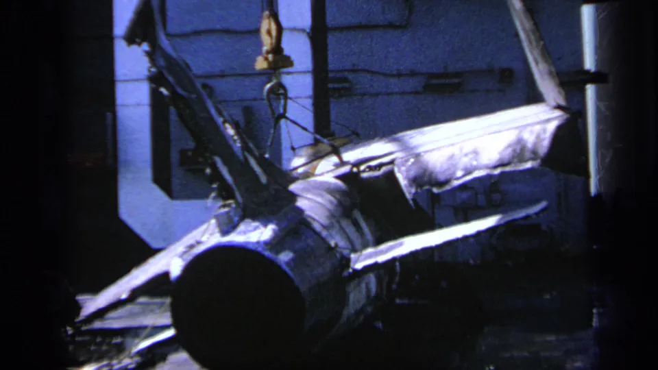 Blue-toned 8mm frame from Greece in 1963 showing a large mechanical piece below visible hoist rigging in a workshop.