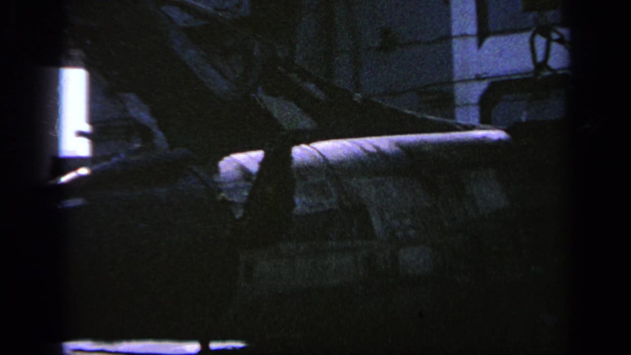 1963 Greece 8mm frame showing part of a camouflaged military helicopter in a dim indoor hangar-like space.