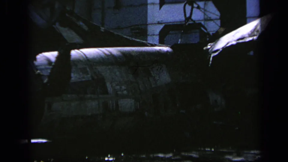 Low-light 8mm view of a camouflaged military helicopter section, partially obscured by hangar shadows in Greece.
