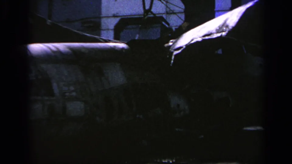 1963 Greece 8mm closeup with a camouflaged helicopter body, dark hangar background, and a bright nearby surface.