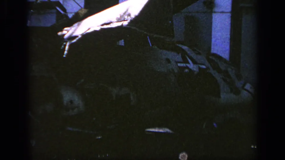 Low-light 8mm frame from Greece showing a bright hand or panel entering near a camouflaged military helicopter.
