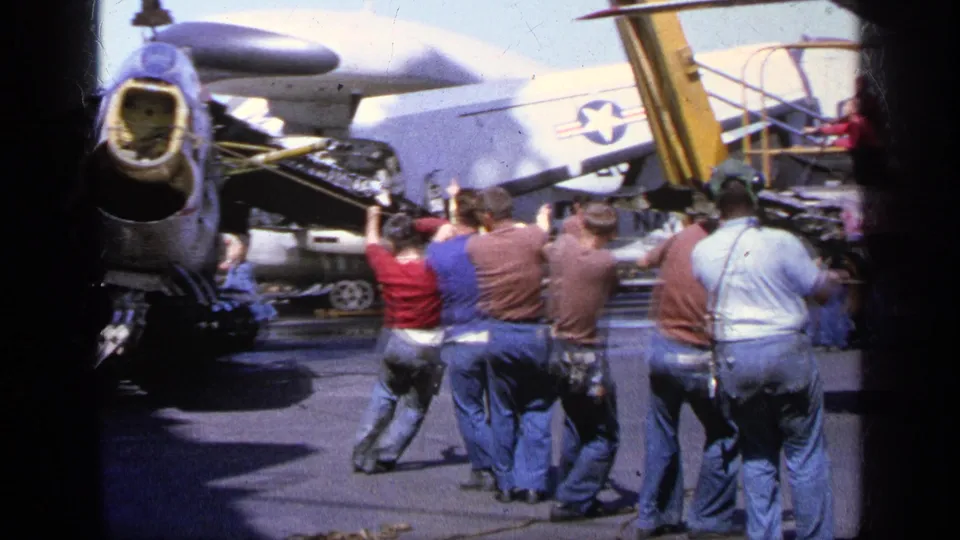 Men walk in a line across a Greek airfield in 1963, with military aircraft sections, a star-marked tail, and service equipment behind them.