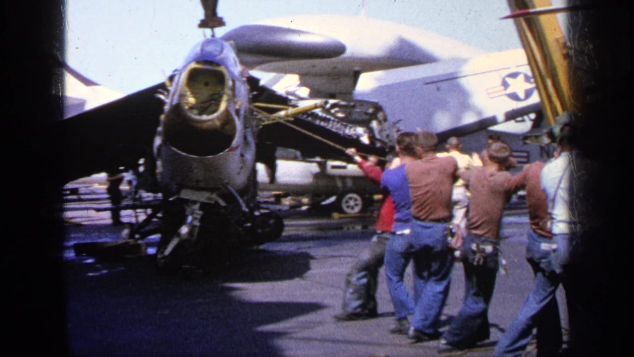 1963 Greece home-movie frame showing crew working near a military aircraft on a carrier deck while a crane line is positioned above.