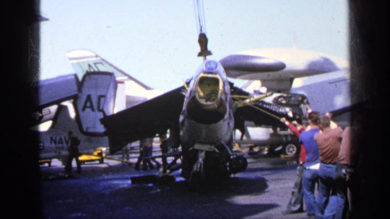 A military aircraft sits on a carrier deck with crew nearby and a crane hook above the nose area.