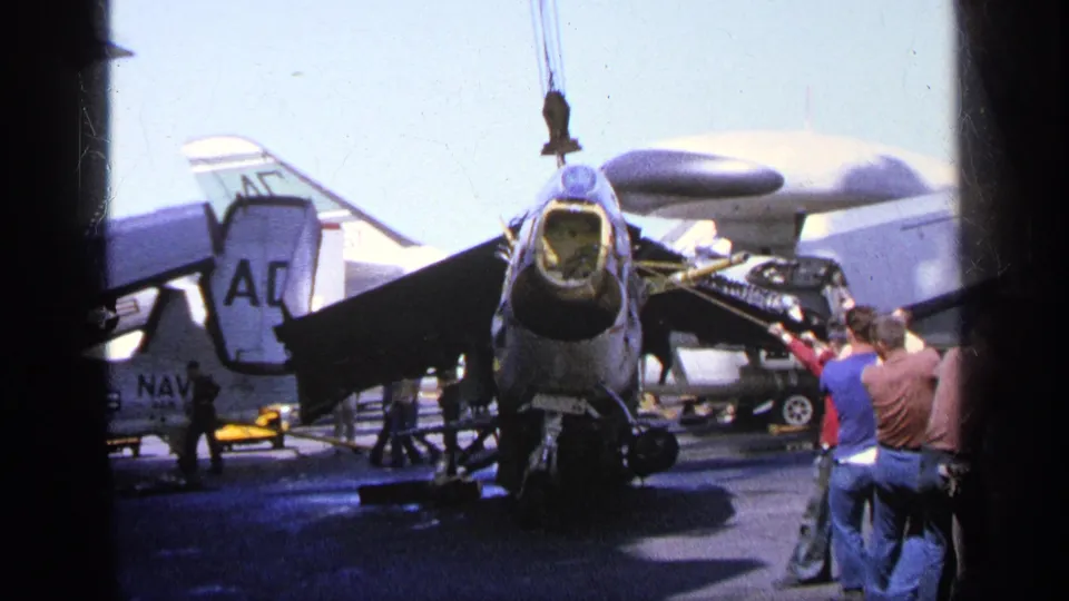 1963 Greece footage frame showing a Navy-marked aircraft on a carrier deck, with crew and lifting gear visible around it.
