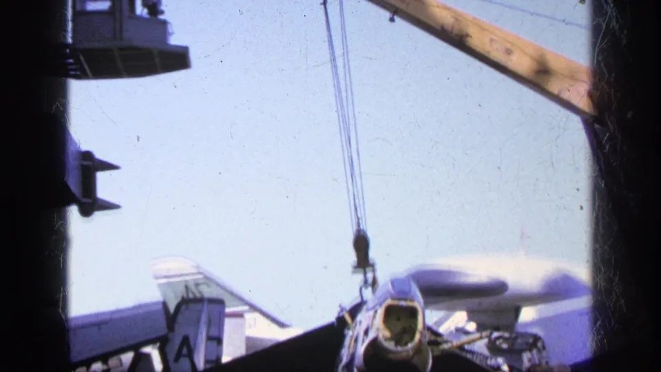 1963 Greece home-movie frame with the aircraft partly obscured by framing, showing a crane line and carrier-deck work area.