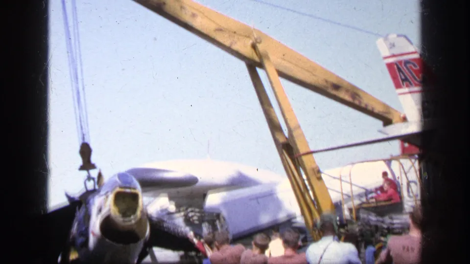 1963 Greece frame showing a crane boom over a Navy-marked aircraft as crew members stand along the carrier deck.
