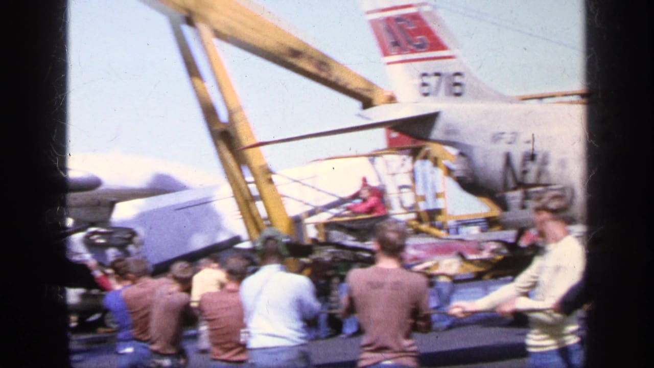 In 1963 Greece, spectators stand close to a U.S. Navy aircraft as a yellow crane and ground activity appear around the tail section.