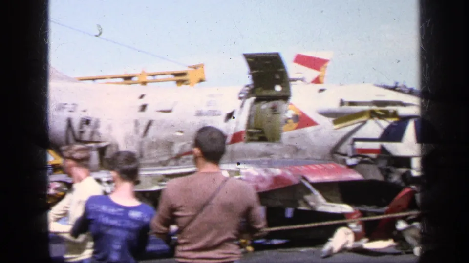 Spectators in 1960s clothing view a U.S. Navy jet in Greece, with an open canopy, wing stores, and military markings visible.