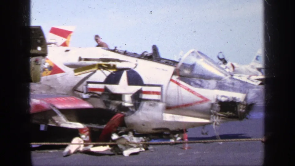 A U.S. Navy aircraft sits on the airfield in Greece with its canopy open, tail markings visible, and ground equipment nearby.