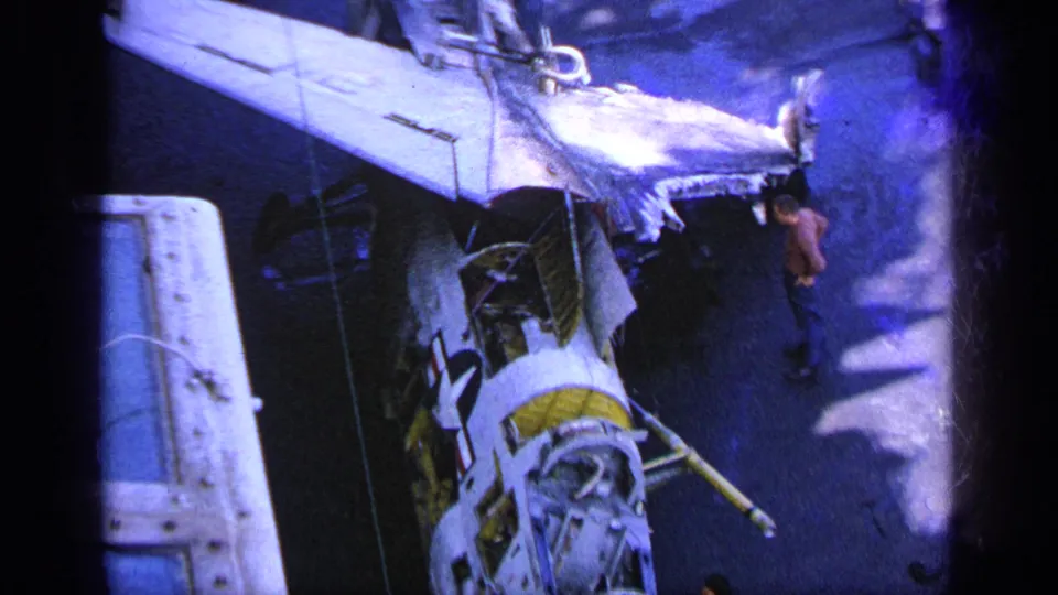 High-angle 1963 Greece frame showing the aircraft wing and damaged fuselage beside crew on the carrier deck.