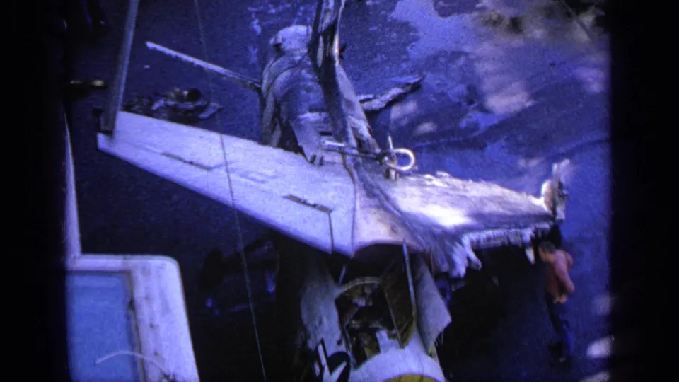 1963 Greece 8mm frame with a torn aircraft wing in the foreground and people standing on the carrier deck.