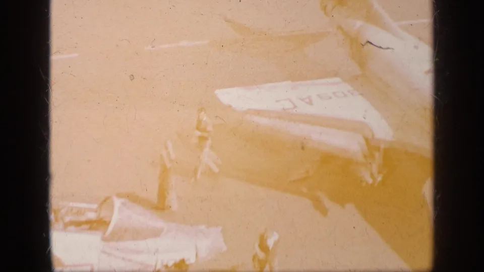 Warmly faded 1963 Greece frame with the jet tail, partial cockpit area, and ground crew figures visible on the airfield surface.