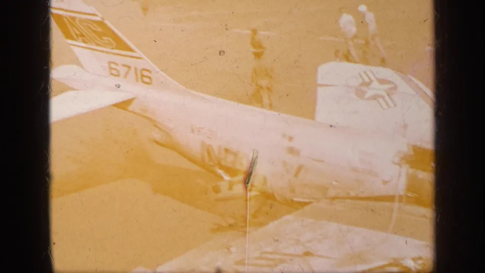 1963 Greece home-movie frame showing a yellow-faded military aircraft on the ground with tail numbers, a star insignia, wing area, and distant personnel.