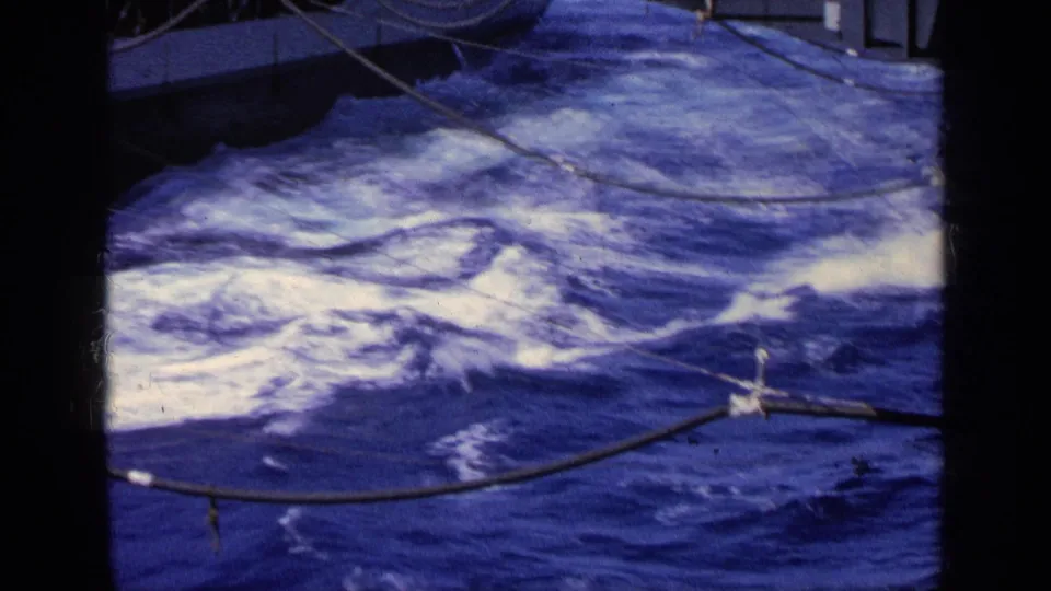 1963 Greece home-movie frame of churning sea beside a cargo ship, with deck cables crossing the foreground and hull partly visible.