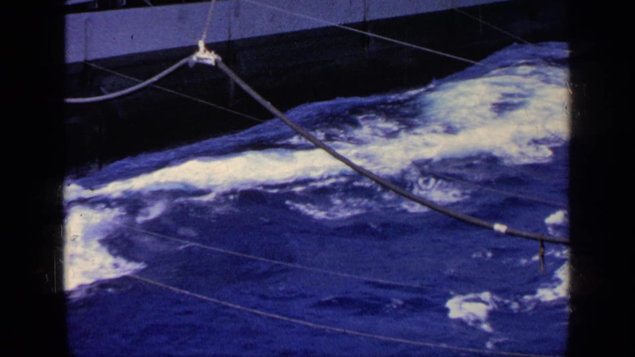 Foamy waves move past the side of a boat with cables and deck hardware overhead.