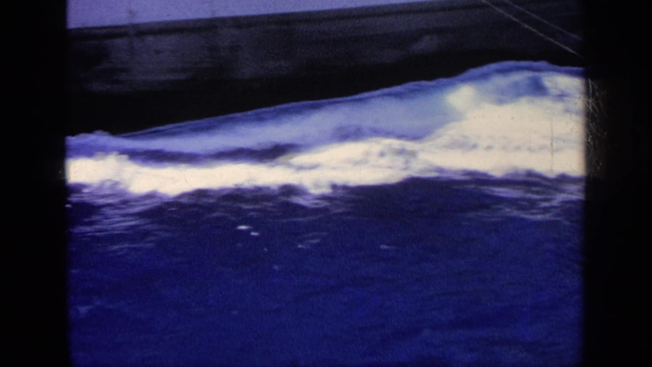 1963 Greece 8mm frame showing dark ship hull edges above blue water with foamy wake and visible film borders.