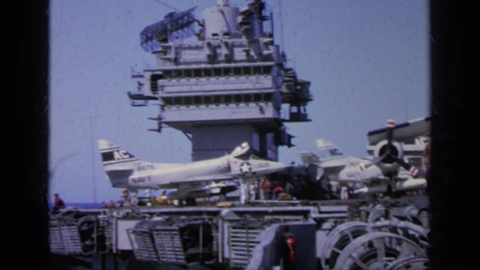 1963 Greece 8mm frame showing a U.S. Navy aircraft carrier deck with parked jet aircraft, crew figures, and radar structures above.