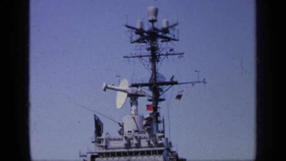 1963 Greece 8mm view focused on the aircraft carrier island mast, radar dishes, antennas, and signal flags against blue sky.
