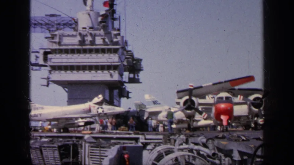 1963 Greece carrier deck showing parked naval aircraft, crew figures, radar structures, and a red-nosed propeller plane.