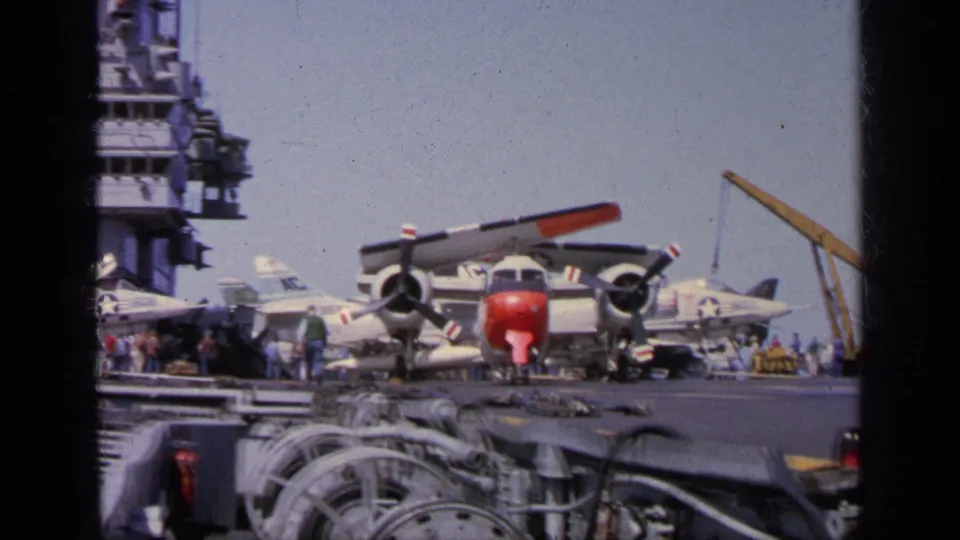 1963 Greece home-movie frame of a carrier flight deck with naval aircraft, crew, deck machinery, and a yellow crane.