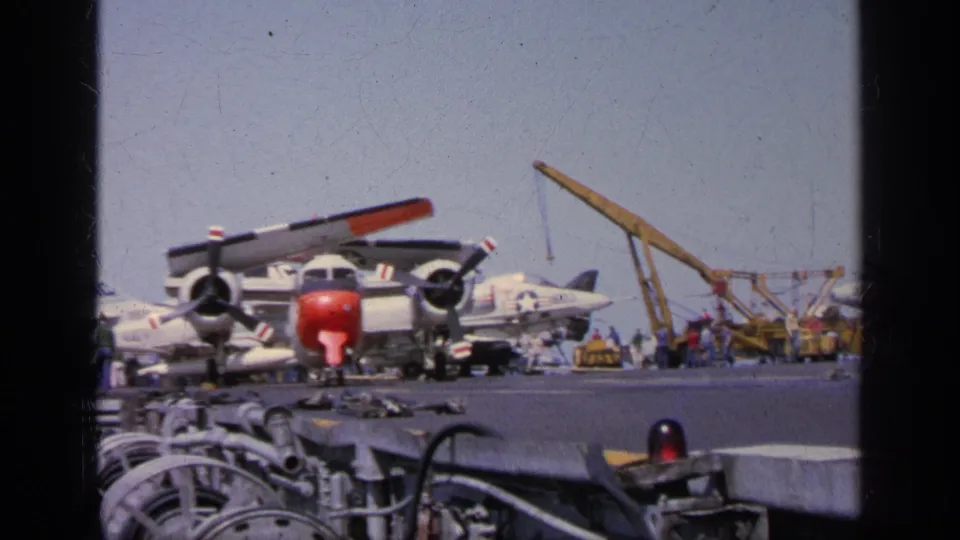 1963 Greece carrier deck view centered on a red-nosed propeller aircraft with a yellow crane and parked jets behind.