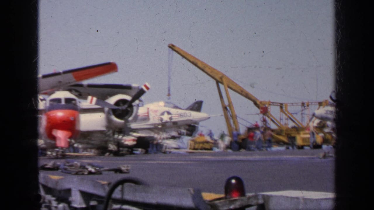 1963 Greece 8mm frame showing Navy personnel near a propeller aircraft, jet fighter, crane, and deck support equipment.