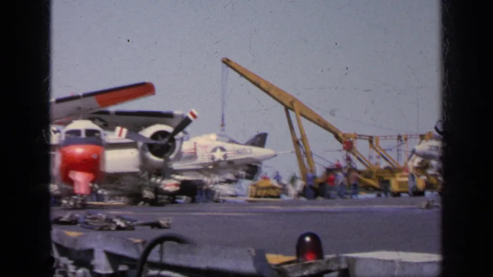 1963 Greece 8mm frame showing Navy personnel near a propeller aircraft, jet fighter, crane, and deck support equipment.
