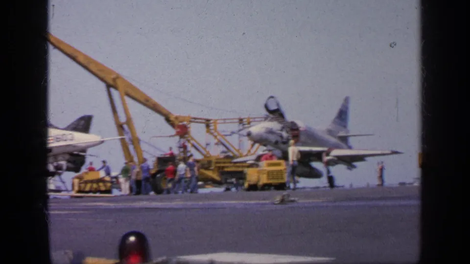 1963 Greece carrier-deck view with a jet aircraft, yellow cranes, support carts, and personnel working nearby.
