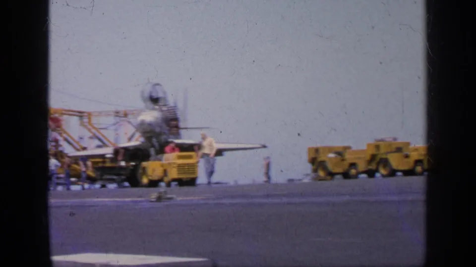 1963 Greece frame showing a jet aircraft at the edge of view, personnel nearby, and yellow tow vehicles crossing the carrier deck.