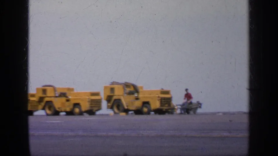 1963 Greece home-movie frame showing yellow support vehicles and a small crew figure on a military flight deck or tarmac.