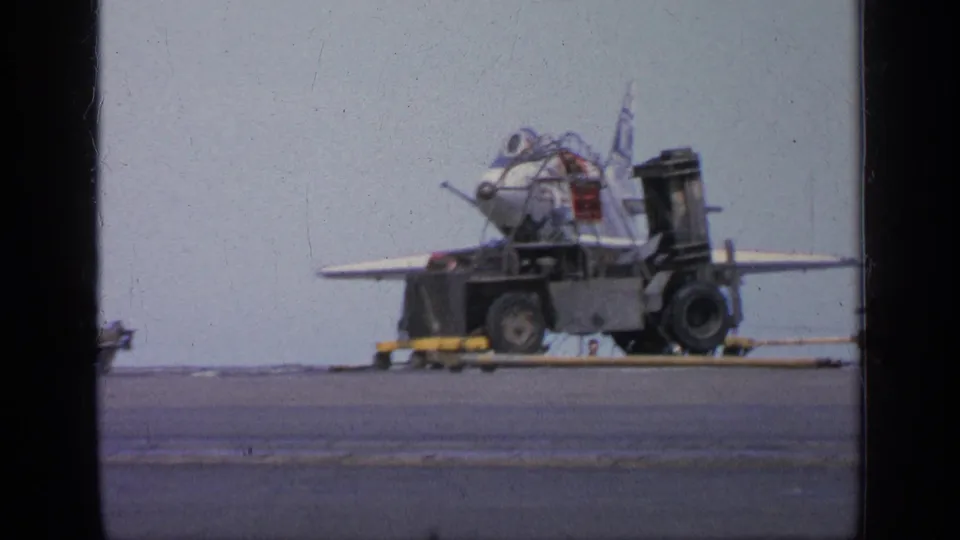 1963 Greece 8mm frame showing a jet aircraft near ground equipment and yellow support vehicles on a military deck or tarmac.
