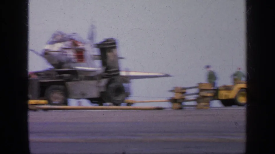 Motion-blurred 1963 Greece frame showing a jet and yellow support vehicles moving or being filmed from a moving viewpoint.