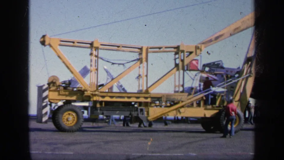 1963 Greece home-movie frame with a large yellow crane or gantry, aircraft equipment, and crew on a military deck or tarmac.