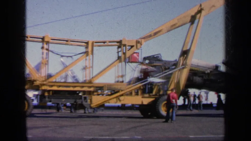 1963 Greece 8mm frame showing a yellow deck crane positioned near a damaged military aircraft while people stand on the carrier deck.
